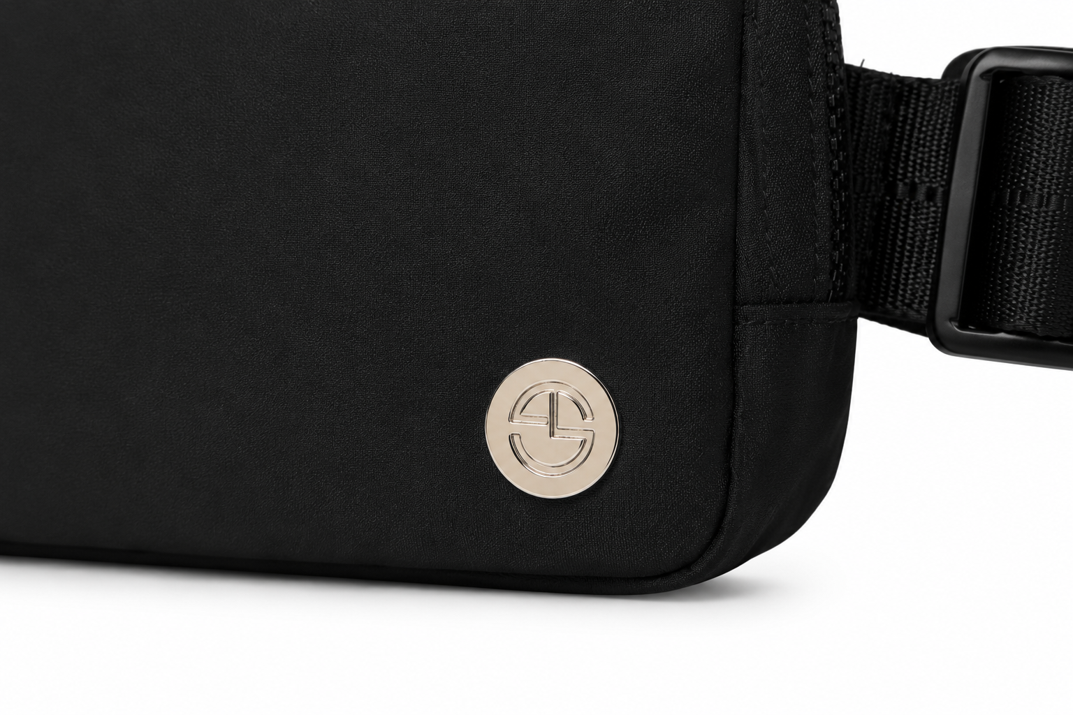 Close-up of a black fabric crossbody bag with a round metallic logo emblem and a black adjustable strap on a white background.
