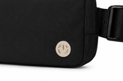 Close-up of a black fabric crossbody bag with a round metallic logo emblem and a black adjustable strap on a white background.
