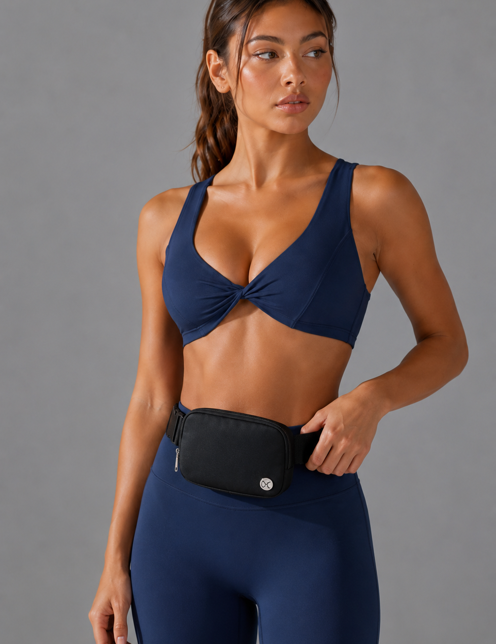 A woman modeling a navy blue athletic sports bra with a front twist detail and matching navy leggings, wearing a black waist bag. She is photographed against a solid gray studio background.