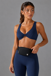 A woman modeling a navy blue athletic sports bra with a front twist detail and matching navy leggings, wearing a black waist bag. She is photographed against a solid gray studio background.